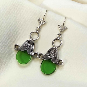Puede incluir: Un par de pendientes colgantes plateados con un diseño geométrico. Cada pendiente presenta una piedra de vidrio verde en forma de lágrima. Los pendientes tienen una parte superior decorativa con un pequeño triángulo y un acento circular.
