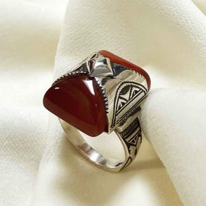 Può includere: Un anello d'argento con una grande pietra di corniola rossa incastonata in un design geometrico. L'anello è finemente dettagliato con una fascia d'argento.