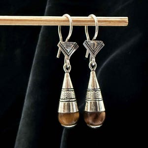 Puede incluir: Un par de pendientes colgantes de plata con gotas de piedra ojo de tigre marrón. Los pendientes tienen un diseño geométrico en la parte superior y una banda de plata texturizada alrededor de la piedra.