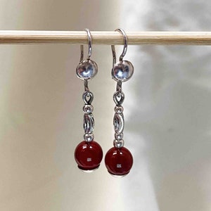 Puede incluir: Un par de pendientes de plata con cuentas de piedras preciosas rojas. Los pendientes tienen una bola de plata en la parte superior y una cadena de cuentas de plata con un diseño decorativo.