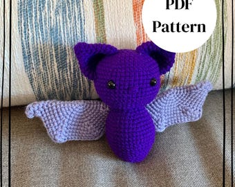 May include: A crocheted bat toy with a deep purple body and head, featuring large pointed ears and light lavender wings. The bat has small black bead eyes and a subtle stitched nose. A white circular graphic displays the text PDF Pattern.