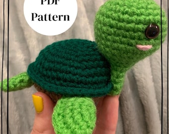 May include: A small crocheted turtle toy featuring a bright green head and flippers, a dark green shell, and a brown underside. The turtle has a single visible dark brown eye and a small pink stitched mouth. A white circular graphic displays the text PDF Pattern.