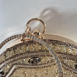 Op de afbeelding: Close-up van een goudkleurige clutch met een decoratief ontwerp. De clutch is versierd met talrijke kleine, sprankelende strass-steentjes en grotere, facetgeslepen edelstenen. Een gebogen handvat met extra strass-steentjes boog over de bovenkant.