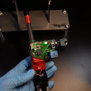 May include: A green and red circuit board with a black and silver key attached. The circuit board is wrapped in red tape and has a green LED light. The text "THE DARK KNIGHT" is visible on a metal surface in the background.