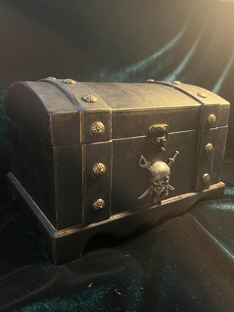Puede incluir: Un cofre del tesoro de madera negra con detalles dorados y un dise&ntilde;o de calavera y huesos cruzados. El cofre tiene una tapa con bisagras y una base decorativa.