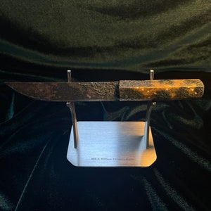 Puede incluir: Un cuchillo antiguo oxidado con mango de madera, expuesto en un soporte plateado. El soporte tiene la inscripción "Bill A. Williams" grabada.