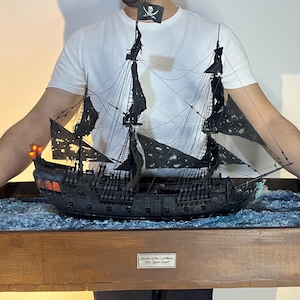 May include: A black model ship of the Black Pearl from the Pirates of the Caribbean movie franchise. The ship is displayed in a wooden box with a blue wave-like surface. The box has a plaque that reads "Pirates of the Caribbean - The Black Pearl."