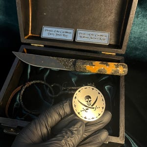 Puede incluir: Una caja de madera con un interior de terciopelo negro. La caja contiene un cuchillo con tem&aacute;tica pirata, una pulsera de cuero y una moneda de plata con un dise&ntilde;o de calavera y huesos cruzados. La caja tiene dos etiquetas, una que dice "Piratas del Caribe Llave de Davy Jones" y la otra que dice "Piratas del Caribe Cuchillo de William Turner".