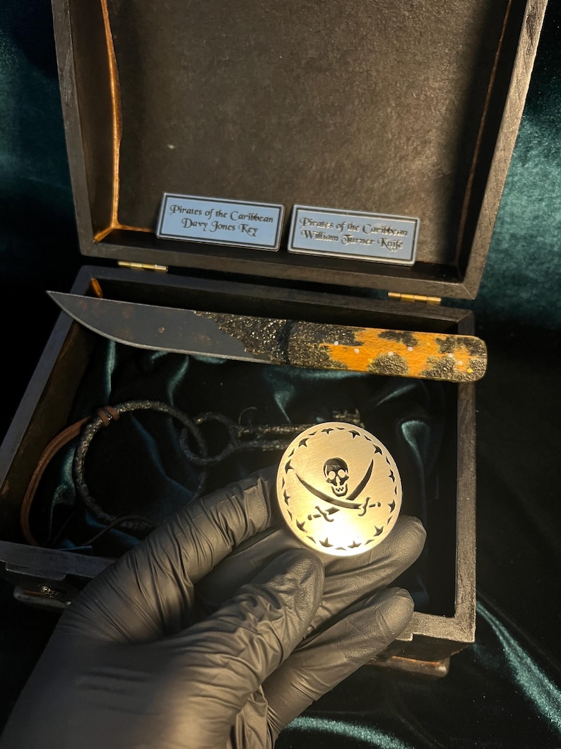 Puede incluir: Una caja de madera con un interior de terciopelo negro. La caja contiene un cuchillo con tem&aacute;tica pirata, una pulsera de cuero y una moneda de plata con un dise&ntilde;o de calavera y huesos cruzados. La caja tiene dos etiquetas, una que dice "Piratas del Caribe Llave de Davy Jones" y la otra que dice "Piratas del Caribe Cuchillo de William Turner".