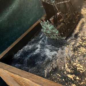 May include: A model of a ship with a black hull and green accents, sinking into a dark blue and white resin sea. The ship is in a wooden frame.