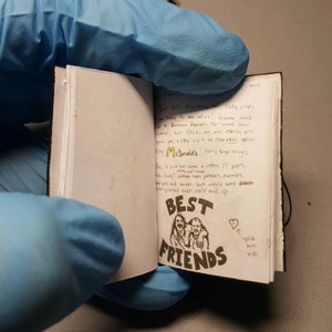 May include: An open notebook with black leather cover and white pages. The pages contain handwritten text in black ink. The text includes the words "Best Friends" and a drawing of two people. The drawing is in black ink and has a heart drawn above it.