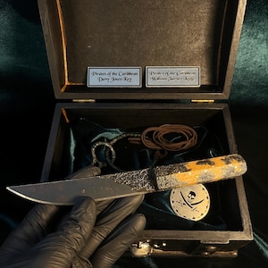 Puede incluir: Una caja de madera negra con un interior de terciopelo verde. La caja contiene un peque&ntilde;o cuchillo negro con mango de madera, un cord&oacute;n de cuero marr&oacute;n y un medall&oacute;n de metal plateado con un dise&ntilde;o de calavera y huesos cruzados. La caja tambi&eacute;n tiene dos placas de metal plateado que dicen "Piratas del Caribe Llave de Davy Jones" y "Piratas del Caribe Cuchillo de William Turner".