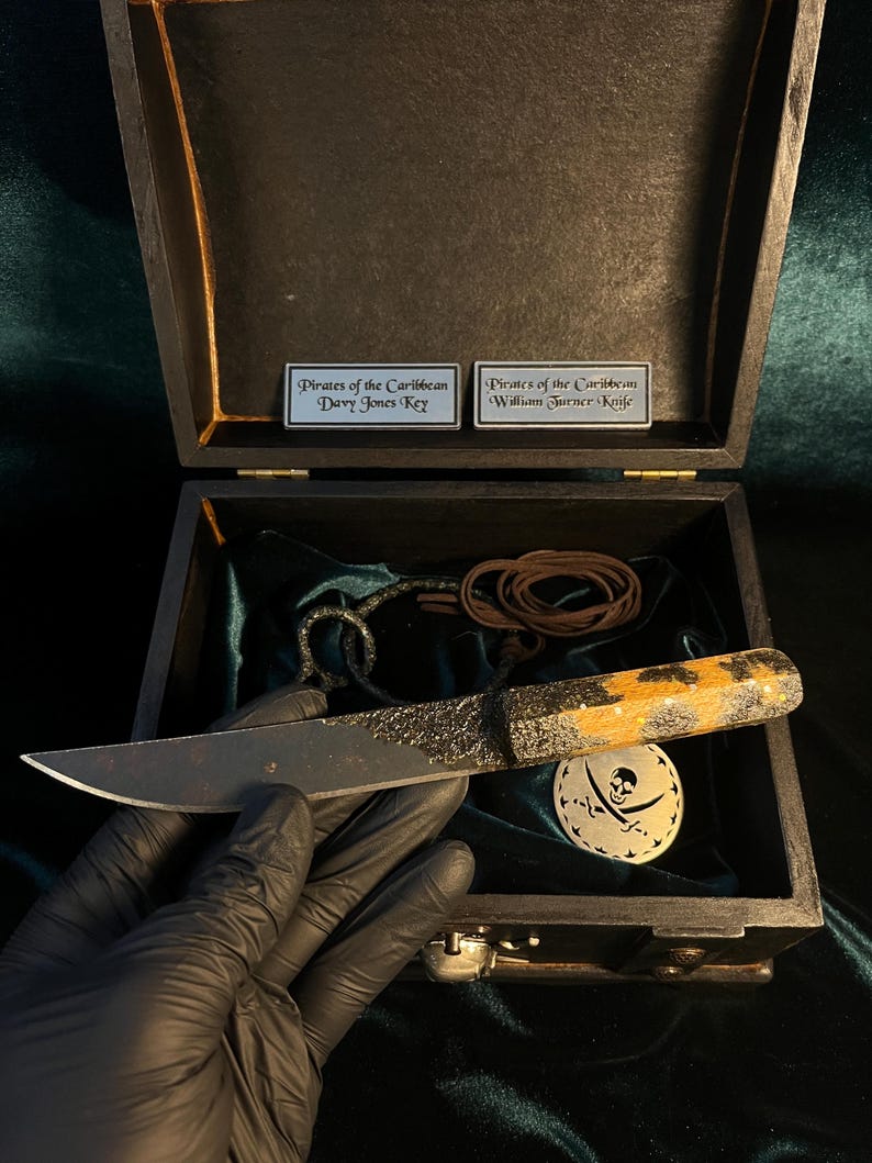 Puede incluir: Una caja de madera negra con un interior de terciopelo verde. La caja contiene un peque&ntilde;o cuchillo negro con mango de madera, un cord&oacute;n de cuero marr&oacute;n y un medall&oacute;n de metal plateado con un dise&ntilde;o de calavera y huesos cruzados. La caja tambi&eacute;n tiene dos placas de metal plateado que dicen "Piratas del Caribe Llave de Davy Jones" y "Piratas del Caribe Cuchillo de William Turner".