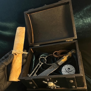 Puede incluir: Un cofre de madera negro con un dise&ntilde;o de calavera y huesos cruzados. El cofre est&aacute; abierto y contiene un rollo de pergamino, una llave, un cuchillo y una correa de cuero.