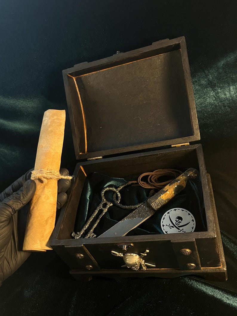 Puede incluir: Un cofre de madera negro con un dise&ntilde;o de calavera y huesos cruzados. El cofre est&aacute; abierto y contiene un rollo de pergamino, una llave, un cuchillo y una correa de cuero.