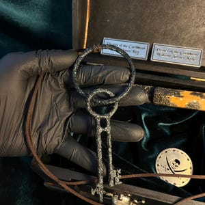 Puede incluir: Una llave de metal negro con una parte superior circular y un cord&oacute;n de cuero marr&oacute;n. La llave est&aacute; en una caja de madera con un dise&ntilde;o de calavera y huesos cruzados. La caja est&aacute; etiquetada "Piratas del Caribe Llave de William Turner".