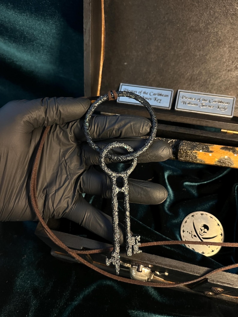Puede incluir: Una llave de metal negro con una parte superior circular y un cord&oacute;n de cuero marr&oacute;n. La llave est&aacute; en una caja de madera con un dise&ntilde;o de calavera y huesos cruzados. La caja est&aacute; etiquetada "Piratas del Caribe Llave de William Turner".