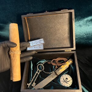 Puede incluir: Un cofre del tesoro de madera con forro de terciopelo verde. El cofre est&aacute; abierto y contiene un rollo enrollado, dos llaves, un cuchillo y un medall&oacute;n de calavera de pirata con huesos cruzados. El cofre est&aacute; etiquetado como "Pirates of the Caribbean Davy Jones Key" y "William Turner - Kelle"