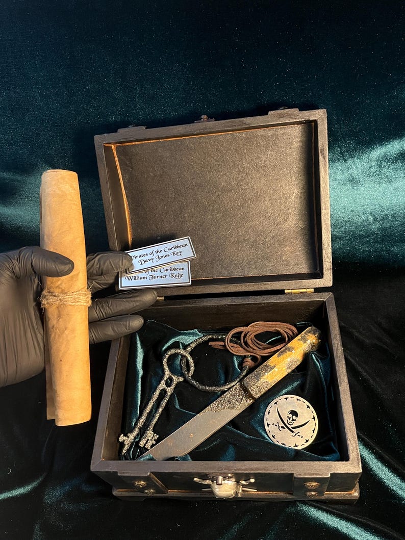 Puede incluir: Un cofre del tesoro de madera con forro de terciopelo verde. El cofre est&aacute; abierto y contiene un rollo enrollado, dos llaves, un cuchillo y un medall&oacute;n de calavera de pirata con huesos cruzados. El cofre est&aacute; etiquetado como "Pirates of the Caribbean Davy Jones Key" y "William Turner - Kelle"