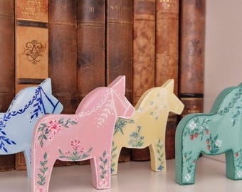 Könnte beinhalten: Vier farbenfrohe Holzpferde, bemalt mit floralen Mustern. Die Pferde sind in Blau, Rosa, Gelb und Grün gehalten und stehen auf einem weißen Regal vor einer Reihe antiker Bücher. Ein charmantes, handgefertigtes Dekorationsstück.