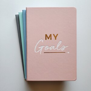 May include: A stack of four notebooks with pink, blue, grey, and light green covers. The top notebook has the words "MY Goals" written in white on the front cover.