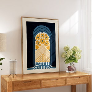May include: A framed print of a colorful abstract design featuring a blue archway with yellow flowers and a geometric pattern. The print is hanging on a white wall above a wooden table.