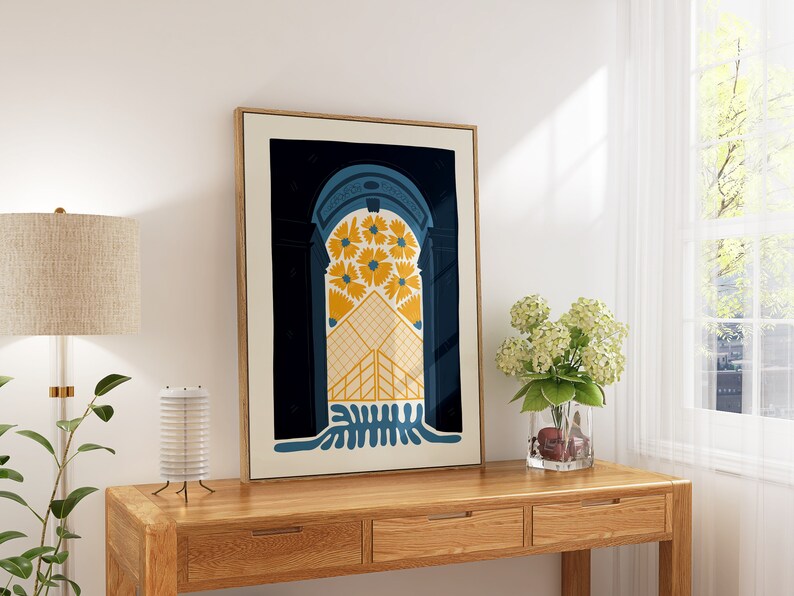 May include: A framed print of a colorful abstract design featuring a blue archway with yellow flowers and a geometric pattern. The print is hanging on a white wall above a wooden table.