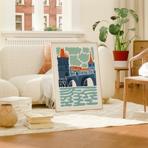 May include: A framed illustration of a bridge with a tower and a church in the background. The illustration is in a modern style with bold colours and simple shapes. The colours are blue, orange, and green.