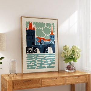 May include: A framed print of a stylised illustration of a bridge with a tower and buildings in blue, red, and orange colours. The bridge is over a body of water with blue and green waves. The background is a cream colour with green clouds.