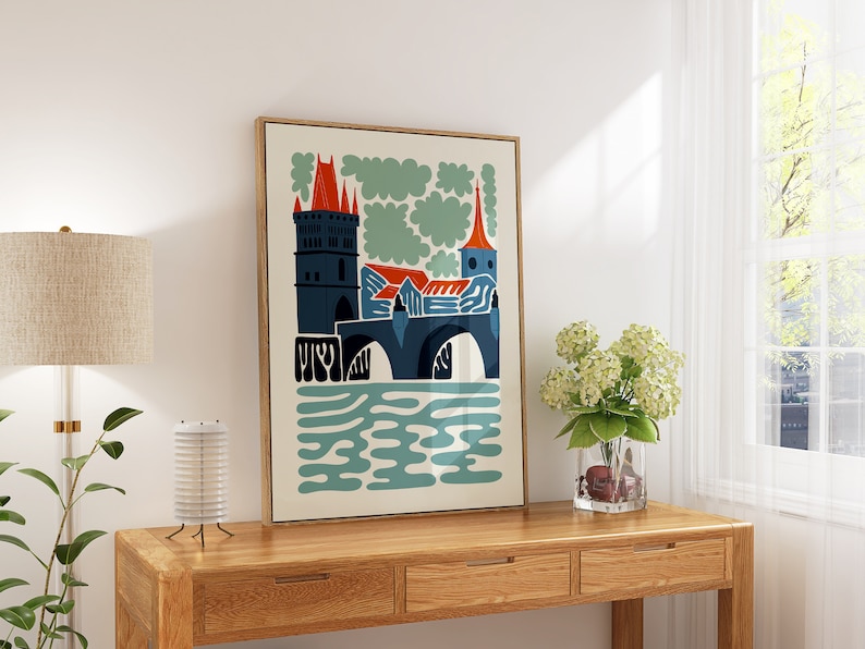May include: A framed print of a stylised illustration of a bridge with a tower and buildings in blue, red, and orange colours. The bridge is over a body of water with blue and green waves. The background is a cream colour with green clouds.