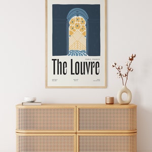 May include: A framed poster with a blue background and a stylized illustration of a doorway with a yellow floral design. The text "The Louvre" is in black and white, with "Paris, France" in smaller text below. The poster is hanging on a white wall above a light brown cabinet with a woven rattan front.