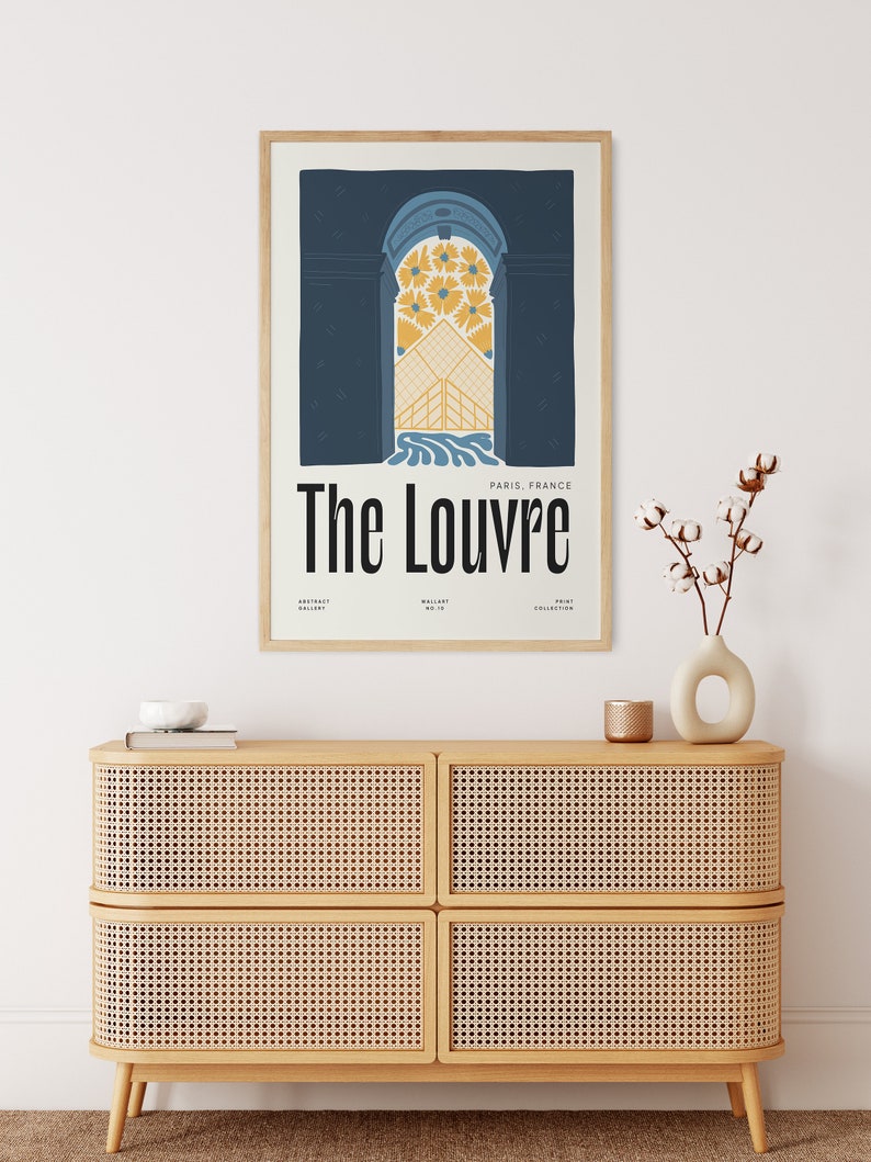 May include: A framed poster with a blue background and a stylized illustration of a doorway with a yellow floral design. The text "The Louvre" is in black and white, with "Paris, France" in smaller text below. The poster is hanging on a white wall above a light brown cabinet with a woven rattan front.