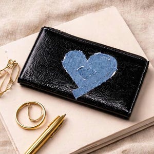 May include: Black patent leather wallet featuring a light blue heart applique. The wallet has a zip closure and is on a notebook with a gold pen and ring. The heart is made of a textured fabric.