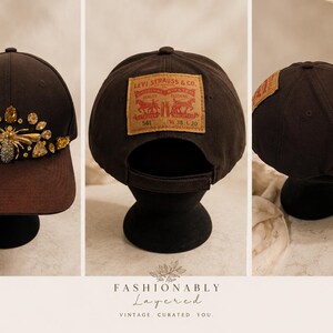 Può includere: Cappellino da baseball marrone scuro con visiera marrone, impreziosito da un'ape gioiello e pietre preziose. La parte posteriore del cappellino presenta una toppa Levi Strauss & Co. Il cappellino è esposto su un supporto nero.