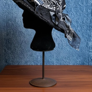 Puede incluir: Un sombrero negro con ala ancha, adornado con un pañuelo bandana negro y blanco con estampado de cachemira. El sombrero presenta una banda de mezclilla y se exhibe sobre una cabeza de maniquí negra sobre un fondo azul.