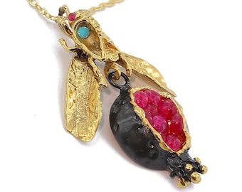 May include: Gold necklace with a pomegranate pendant. The pomegranate is open and filled with red gemstones. A small gold bee with turquoise and red gemstones is attached to the top of the pendant.