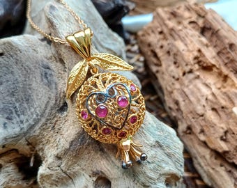 May include: A gold necklace with a pendant shaped like a pomegranate. The pendant is made of filigree metal and has pink gemstones. The pendant is hanging on a wooden surface.