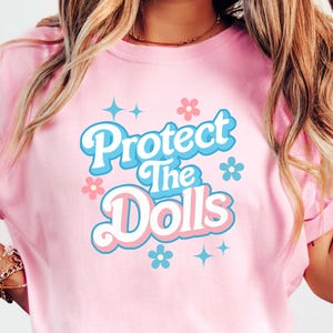 Może przedstawiać: Różowa koszulka z grafiką w stylu retro, na której widnieje napis "Protect the Dolls" w niebiesko-różowych literach z kwiatami i gwiazdami.