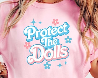 May include: Pink t-shirt with a retro-style graphic that says "Protect the Dolls" in blue and pink lettering with flowers and stars.