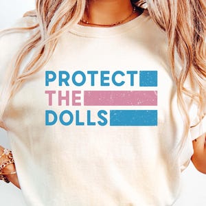 Może przedstawiać: Biała koszulka z napisem "PROTECT THE DOLLS" w niebieskich i różowych literach. Tekst jest w stylu zużytego czcionki.