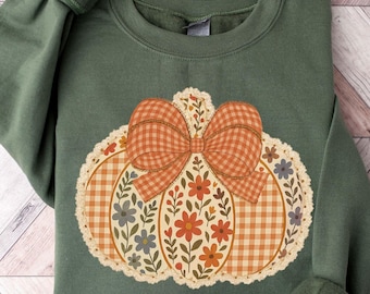 May include: A green sweatshirt featuring a pumpkin design. The pumpkin is decorated with a plaid bow and floral accents in shades of orange, cream, and blue. The sweatshirt has long sleeves with ribbed cuffs, perfect for fall.