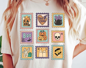 May include: Cream-colored t-shirt featuring a grid of Halloween-themed postage stamp designs. The stamps showcase various spooky illustrations, including a haunted house, pumpkins, and a skull. The shirt is a relaxed fit.
