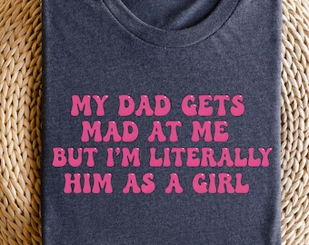 May include: A navy blue t-shirt with the text "MY DAD GETS MAD AT ME BUT I'M LITERALLY HIM AS A GIRL" in pink, retro-style font. The shirt is displayed on a round, woven wicker surface.