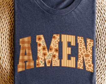 May include: A navy blue t-shirt with the word "AMEN" in a collegiate-style font. The letters feature various patterns, including plaid, floral, stripes, and leopard print with a pumpkin graphic, creating a fall-themed design.