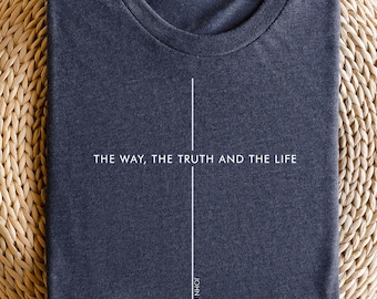 May include: A navy blue t-shirt with the text "THE WAY, THE TRUTH AND THE LIFE" and "JOHN 14:6" in white. The shirt is folded and resting on a woven, circular surface. The t-shirt has a crew neck.