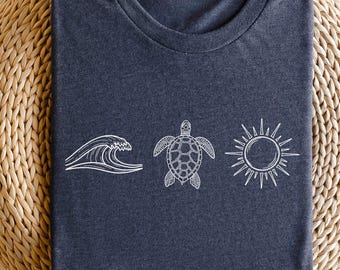 Könnte beinhalten: Ein marineblaues T-Shirt mit einer weißen Grafik einer Welle, einer Meeresschildkröte und einer Sonne. Das T-Shirt ist gefaltet und liegt auf einer gewebten, kreisförmigen Oberfläche. Das Design ist schlicht und erinnert an ein Strandthema.