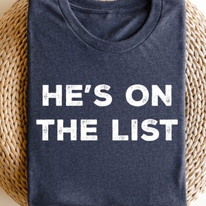 Może przedstawiać: Granatowa koszulka z napisem "HE'S ON THE LIST" w białych literach. Koszulka jest złożona i spoczywa na tkanej, okrągłej powierzchni. Koszulka ma okrągły dekolt.