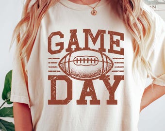 May include: Cream-colored t-shirt with a brown football graphic and the words "GAME DAY." The design includes a football and horizontal lines. The shirt is a casual, short-sleeved style.