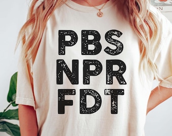 May include: Cream-colored t-shirt with the black text "PBS NPR FDT" in a bold, sans-serif font. The shirt is worn by a person, paired with dark green shorts. A bracelet and necklace are visible.