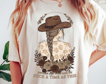 May include: Cream-colored t-shirt featuring a graphic of a woman wearing a cowboy hat, with a long braid, and floral accents. The text "Perhaps you were born for such a time as this" is printed below the graphic.